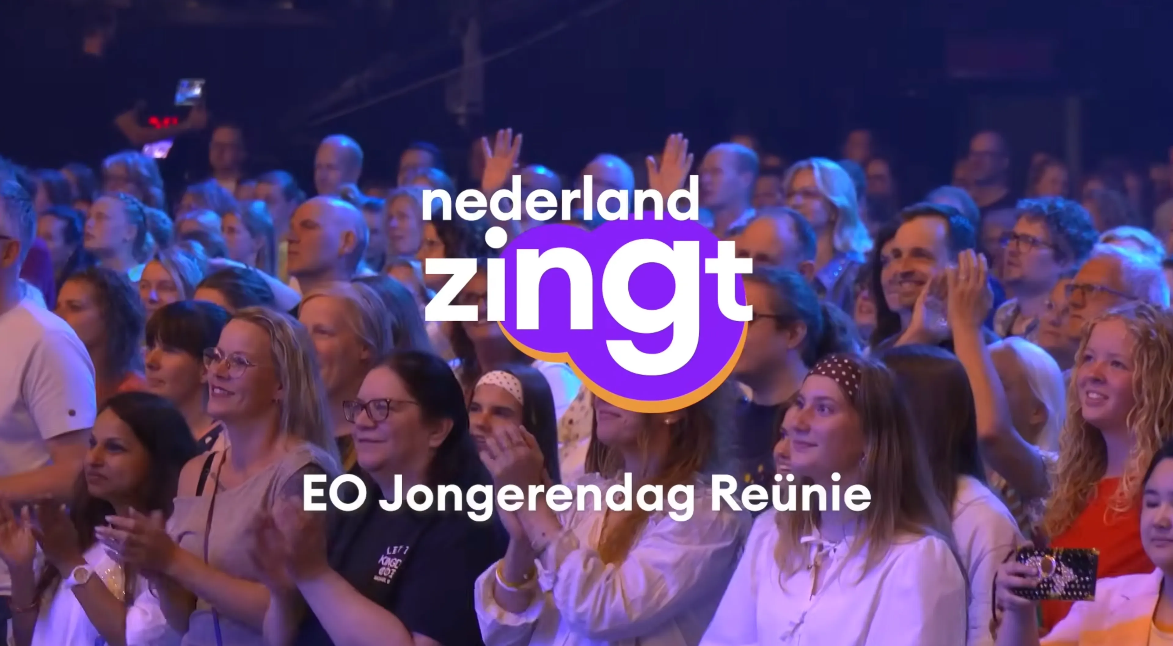 Nederland Zingt in de EO Jongerendag Reünie: een tv-achtige opname van een grote zaal waar een enthousiast publiek naar een podium kijkt, terwijl het programma-logo boven in een paarse badge zichtbaar is en de sfeer wordt versterkt door live-achtige elementen.